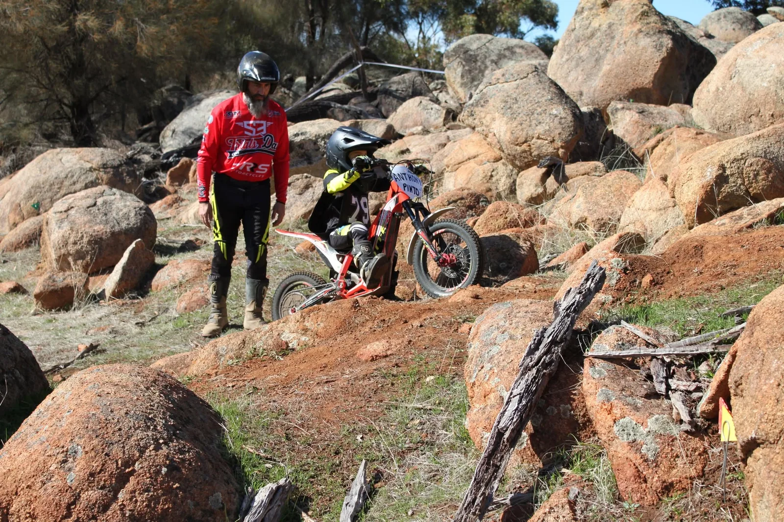 Young rider on a small electric trials bike climbing a rocky section with a mentor nearby
