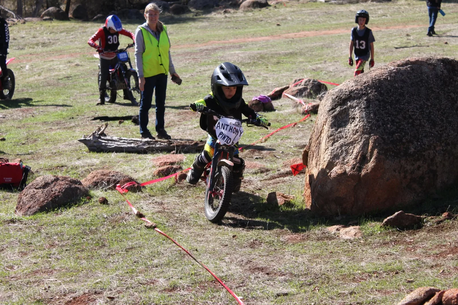 Very young trials rider turning around a rock while adults and other juniors look on